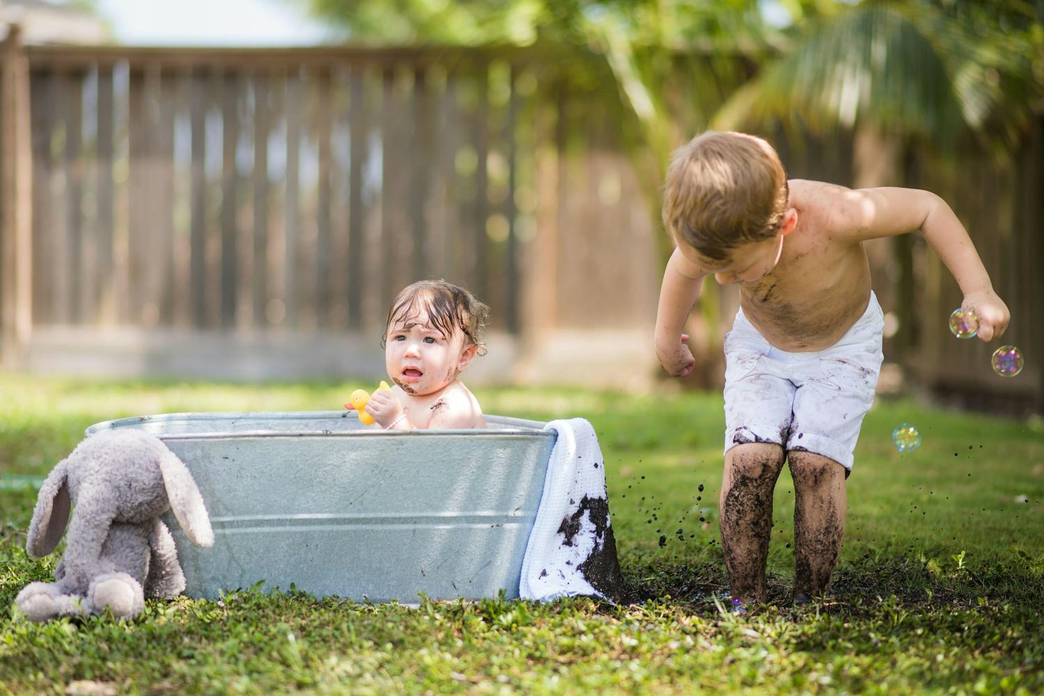 Baby taking a bath in metal tub with brother playing in the mud next to him