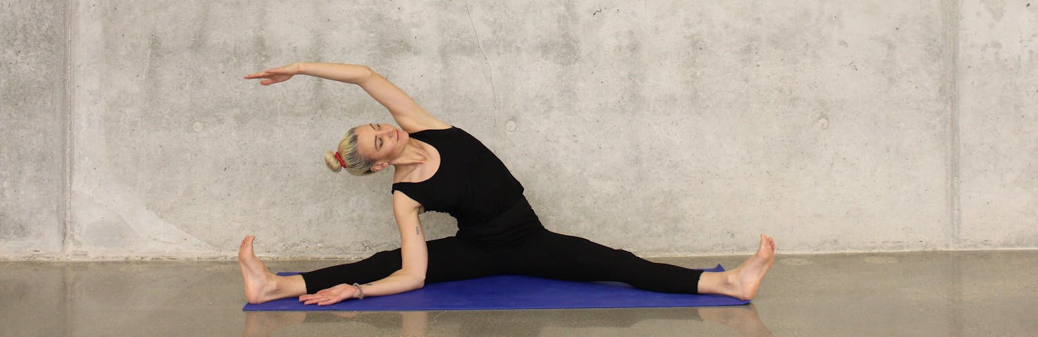 Woman wearing all black against gray wall doing seated side stretch