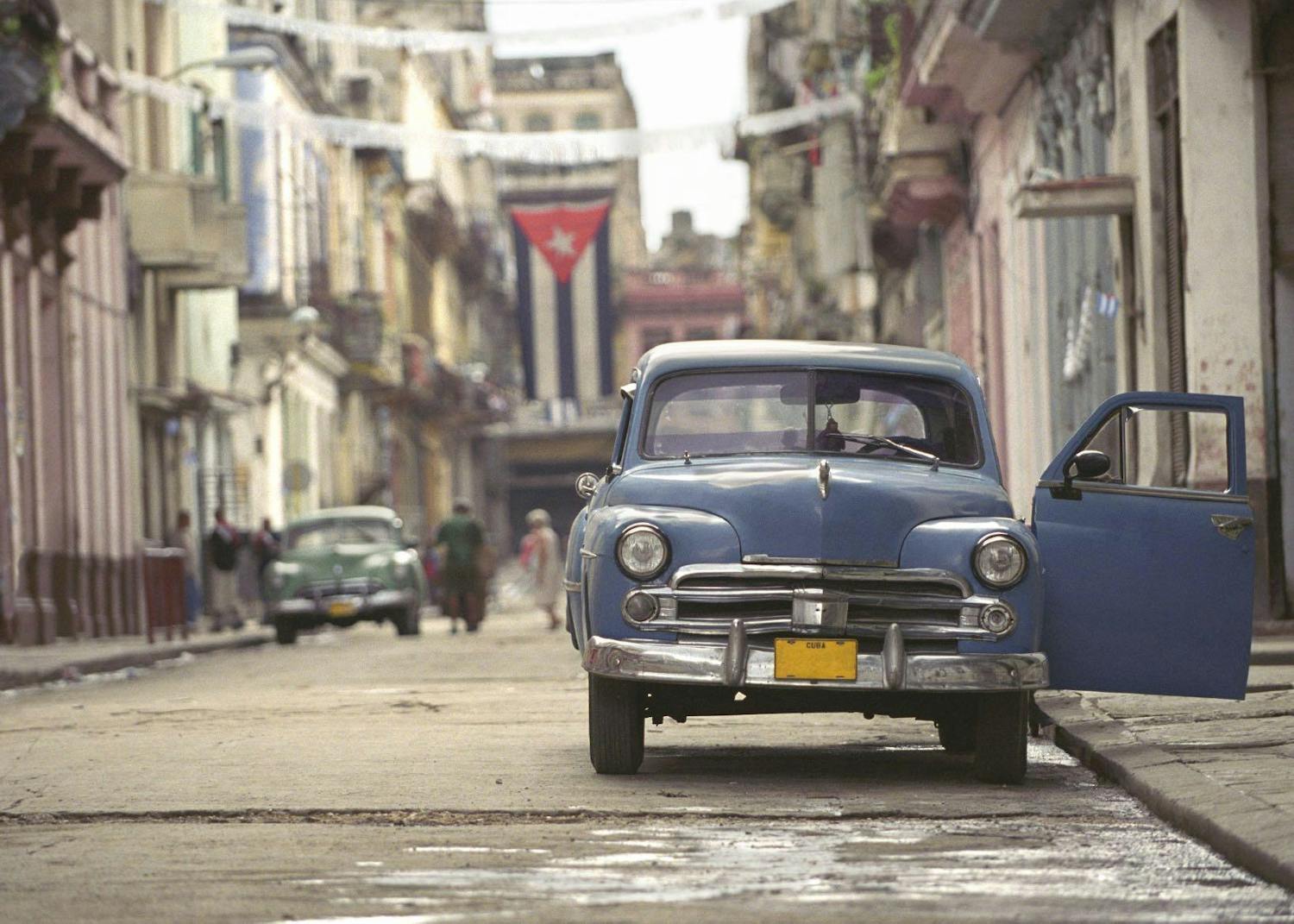 An old blue car parked on an empty street in havana with the cuban flag hanging