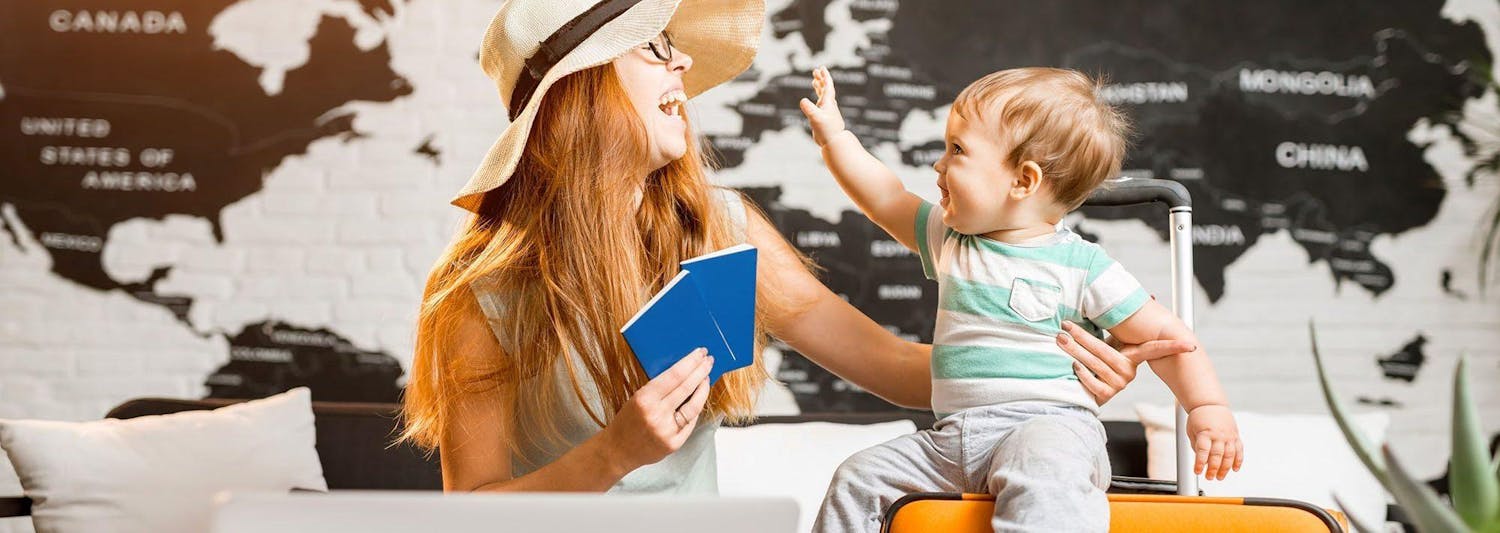Little boy sitting on orange luggage with mother wearing hat and holding passports
