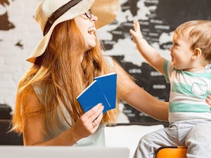Little boy sitting on orange luggage with mother wearing hat and holding passports